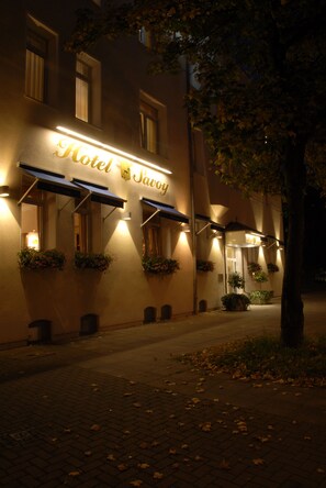 A hotel facade at night with illuminated signage, awnings, and potted plants. - Hotel Savoy Hannover (Hannover)
