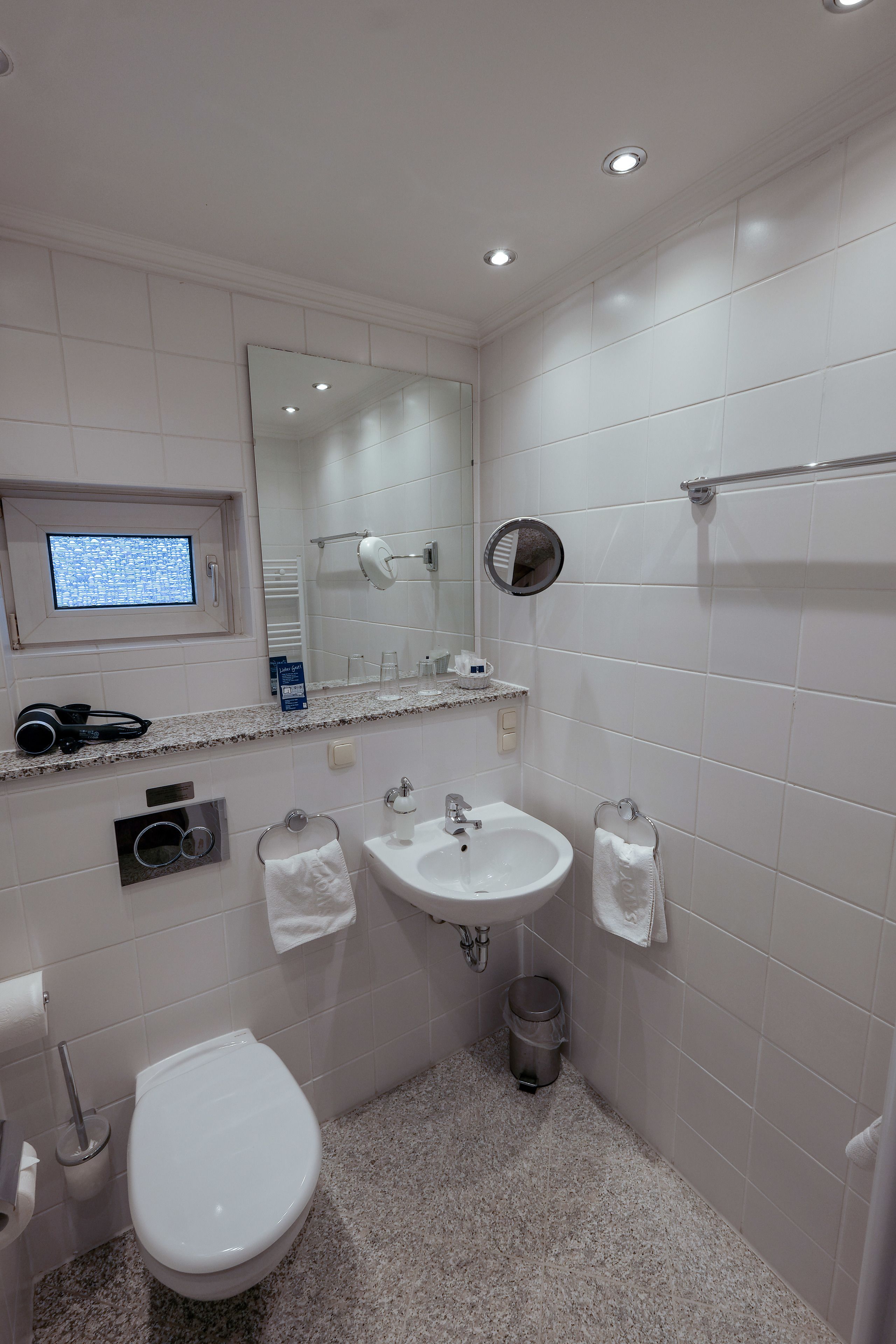 A modern bathroom with a toilet, sink, and mirror. There is a window above the toilet and a small shelf with toiletries.
