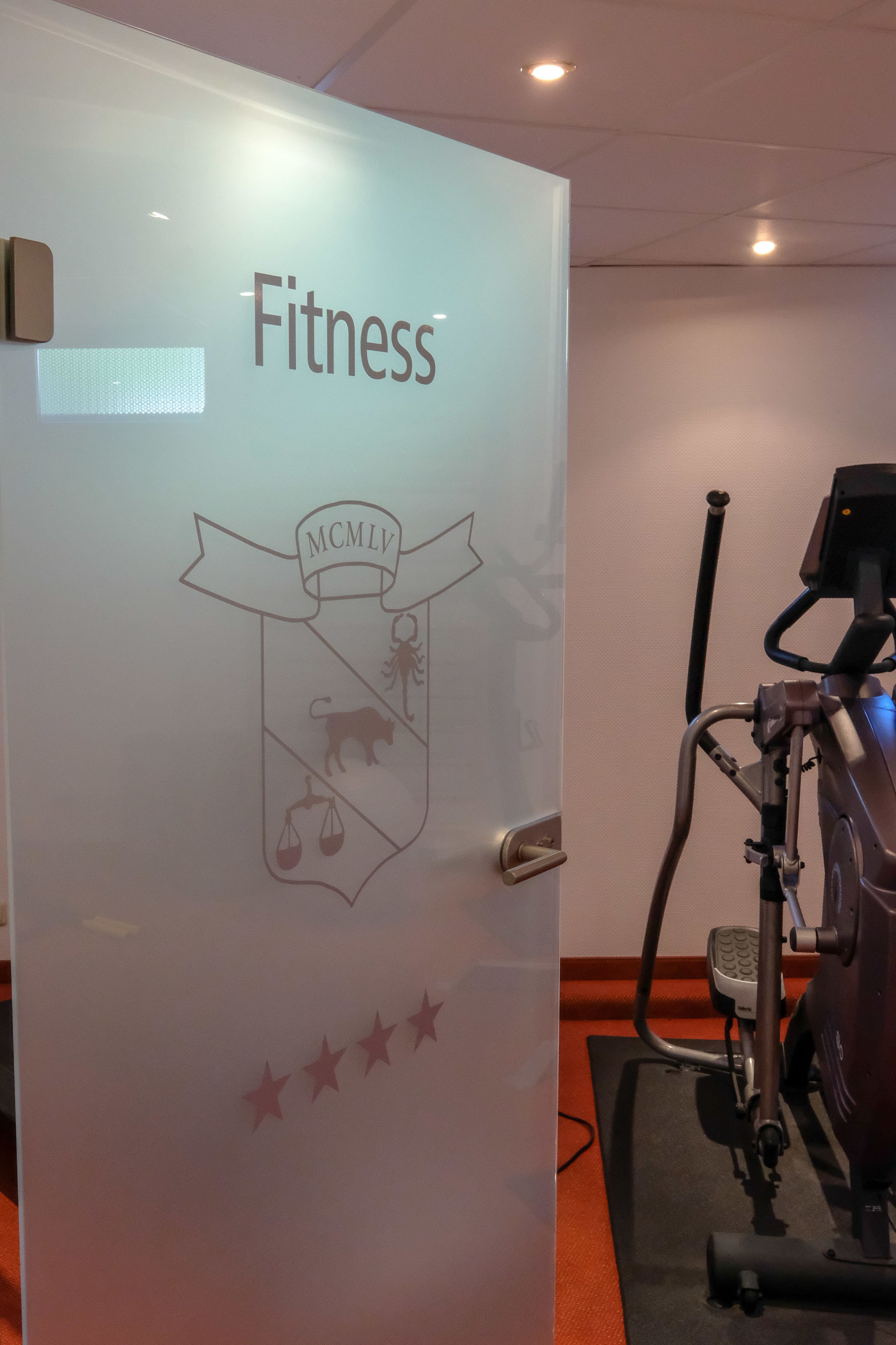 Uma sala de ginástica com uma porta identificada com a palavra 'Fitness' e um logotipo com 'MCMLV' e um brasão.