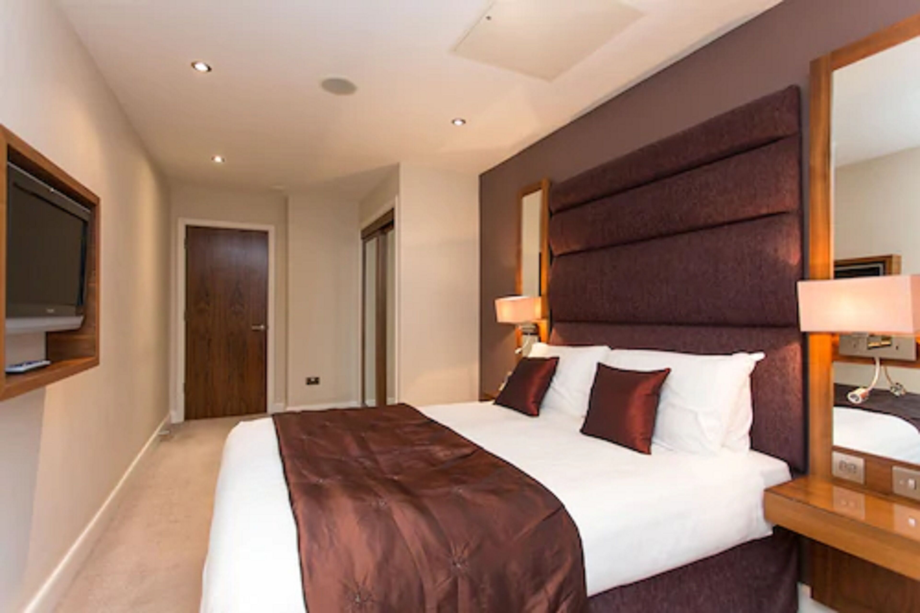A hotel room with a large bed, a wooden headboard, a bedside table with a lamp, and a mirror.
