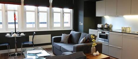 Una sala moderna con un sofá gris, una mesa de centro y una mesa de comedor. Hay una cocina con armarios blancos y una estufa. Amplios ventanales aportan luz natural.