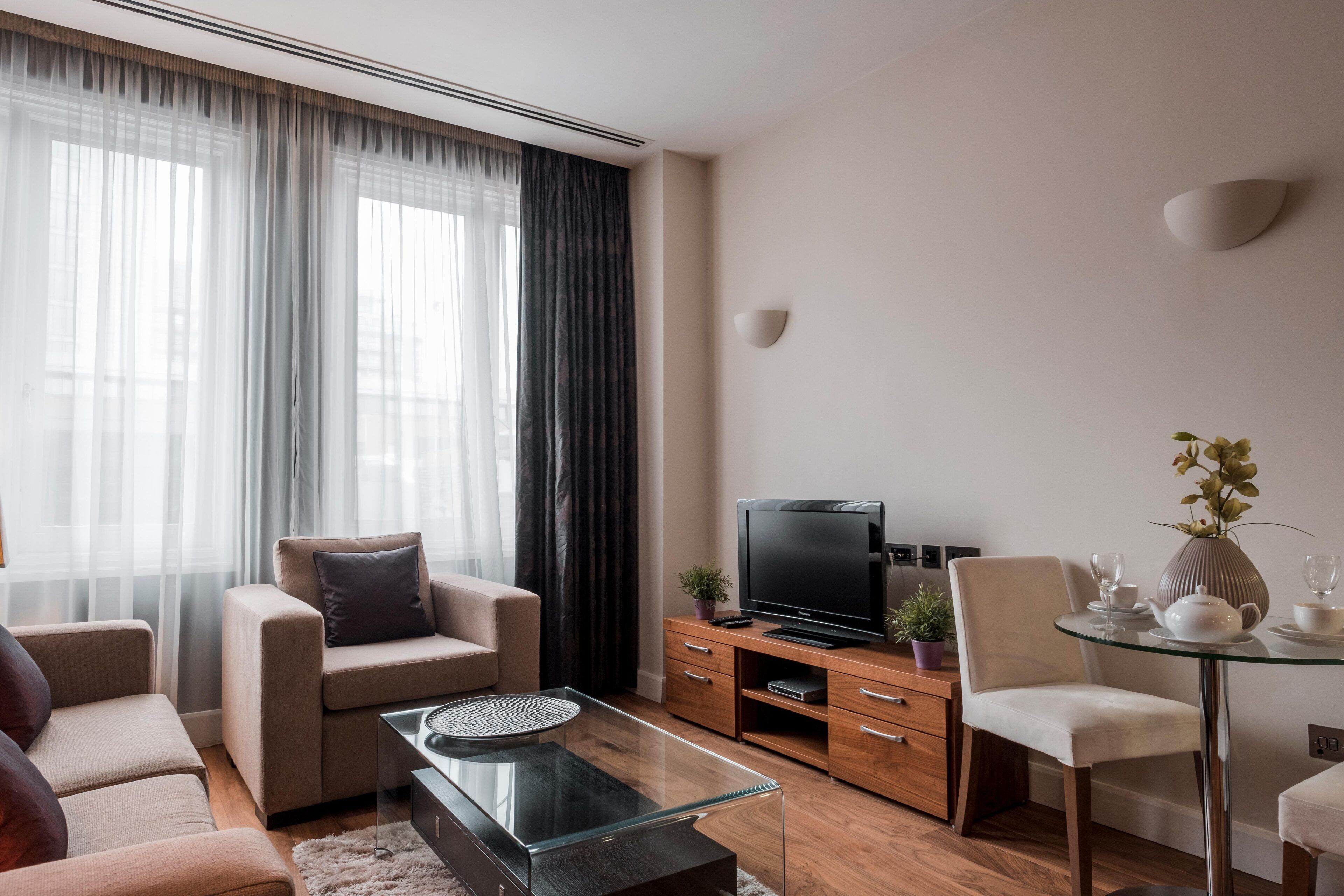 A modern living room with a sofa, a glass coffee table, a TV, and a dining area with a glass table and chairs.