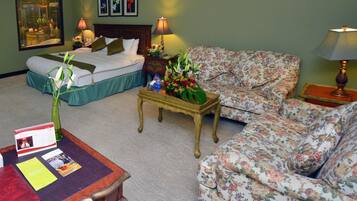 Una habitación de hotel con una cama, dos sofás florales, una pequeña mesa de madera con un jarrón de flores, una mesita de noche con una lámpara y un cuadro en la pared.