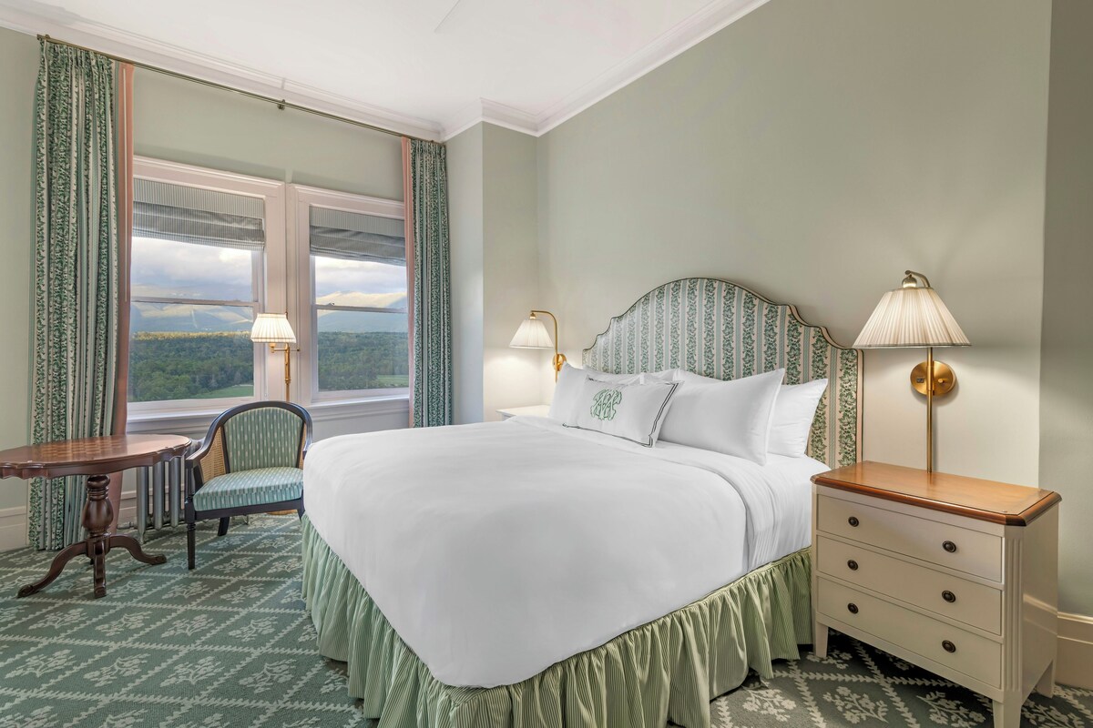 Une chambre d’hôtel avec un grand lit, deux lampes de chevet, une chaise, une petite table et une vue sur les montagnes par la fenêtre.
