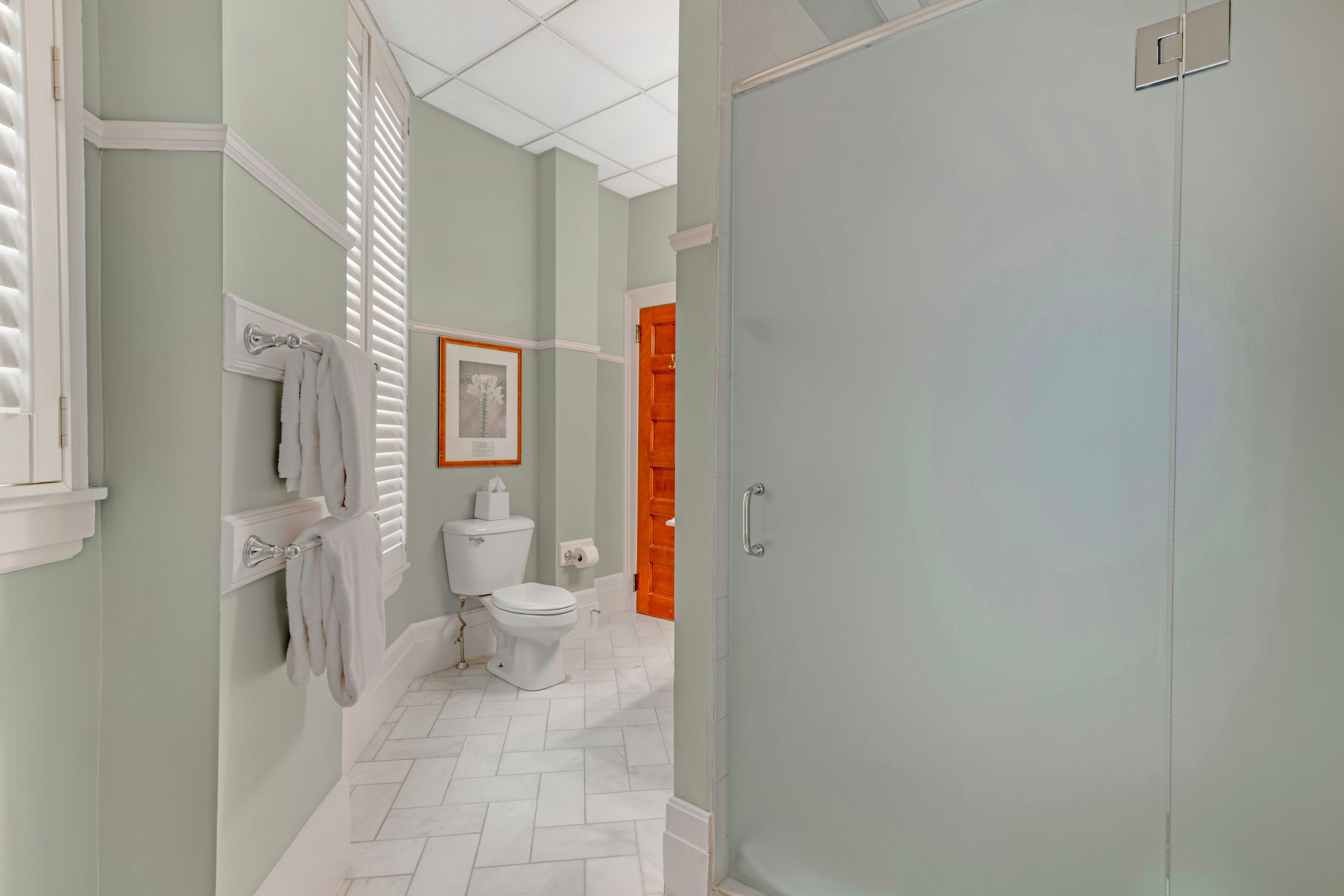 A bathroom with a toilet, a towel rack with a towel, a framed picture on the wall, and a door with a handle.