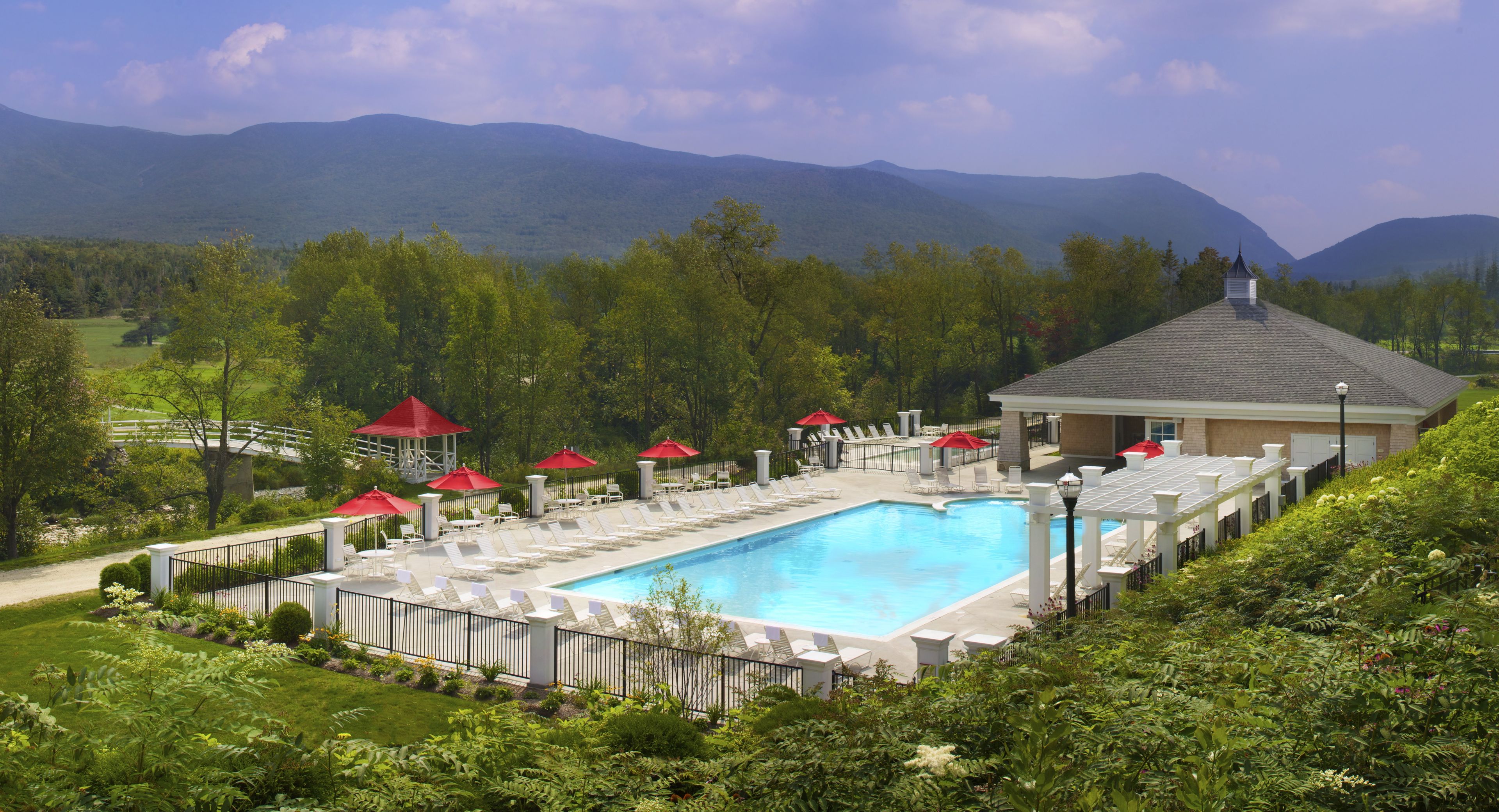 A swimming pool with lounge chairs and umbrellas, surrounded by greenery and mountains in the background.