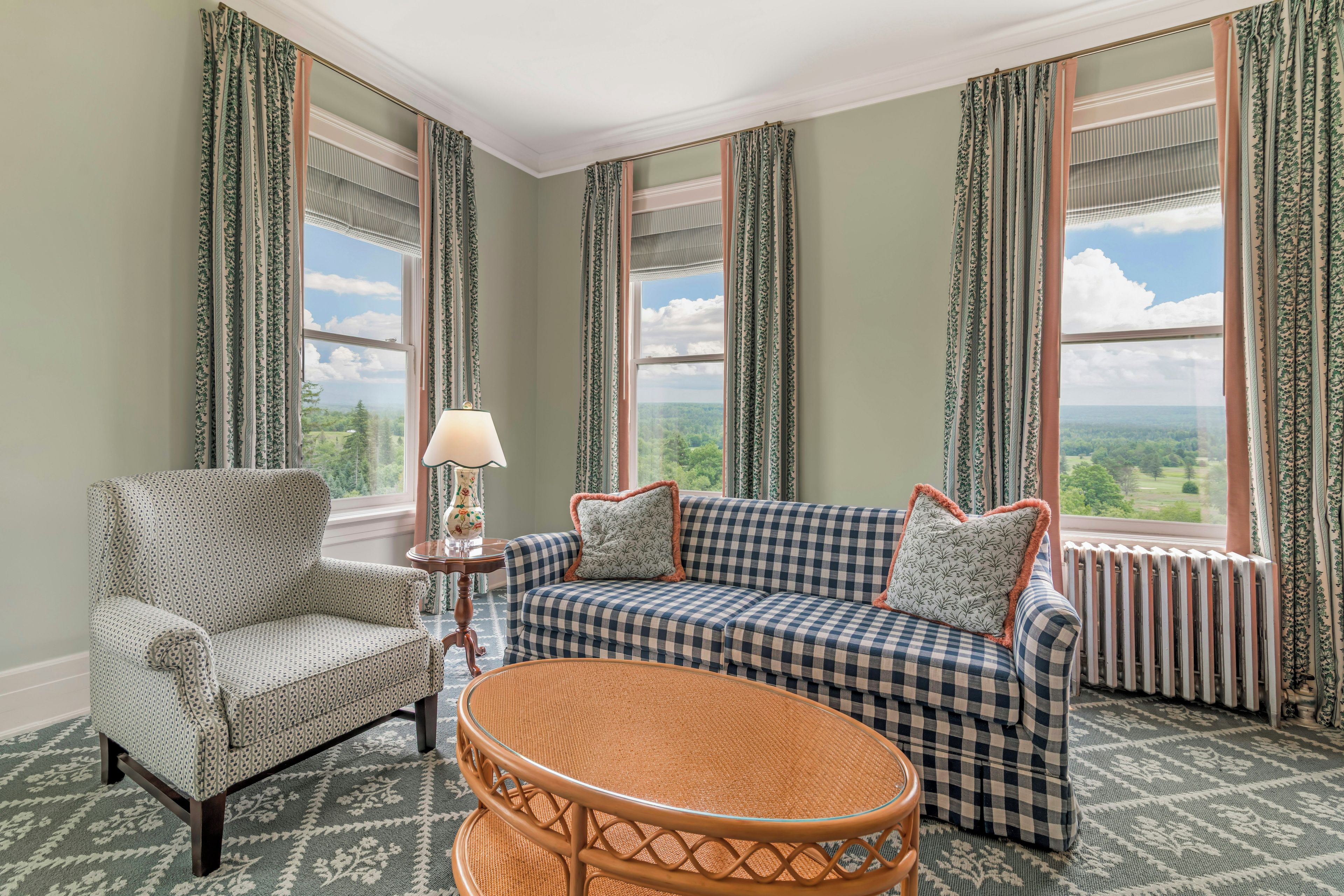 A room with a checkered sofa, a patterned armchair, a wicker table, and a view of the outdoors through large windows.