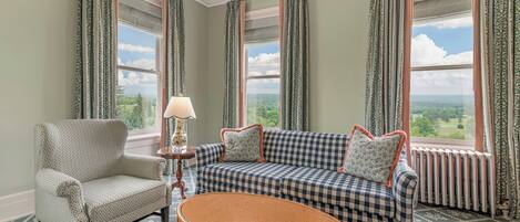A room with a checkered sofa, a patterned armchair, a wicker table, and a view of the outdoors through large windows.