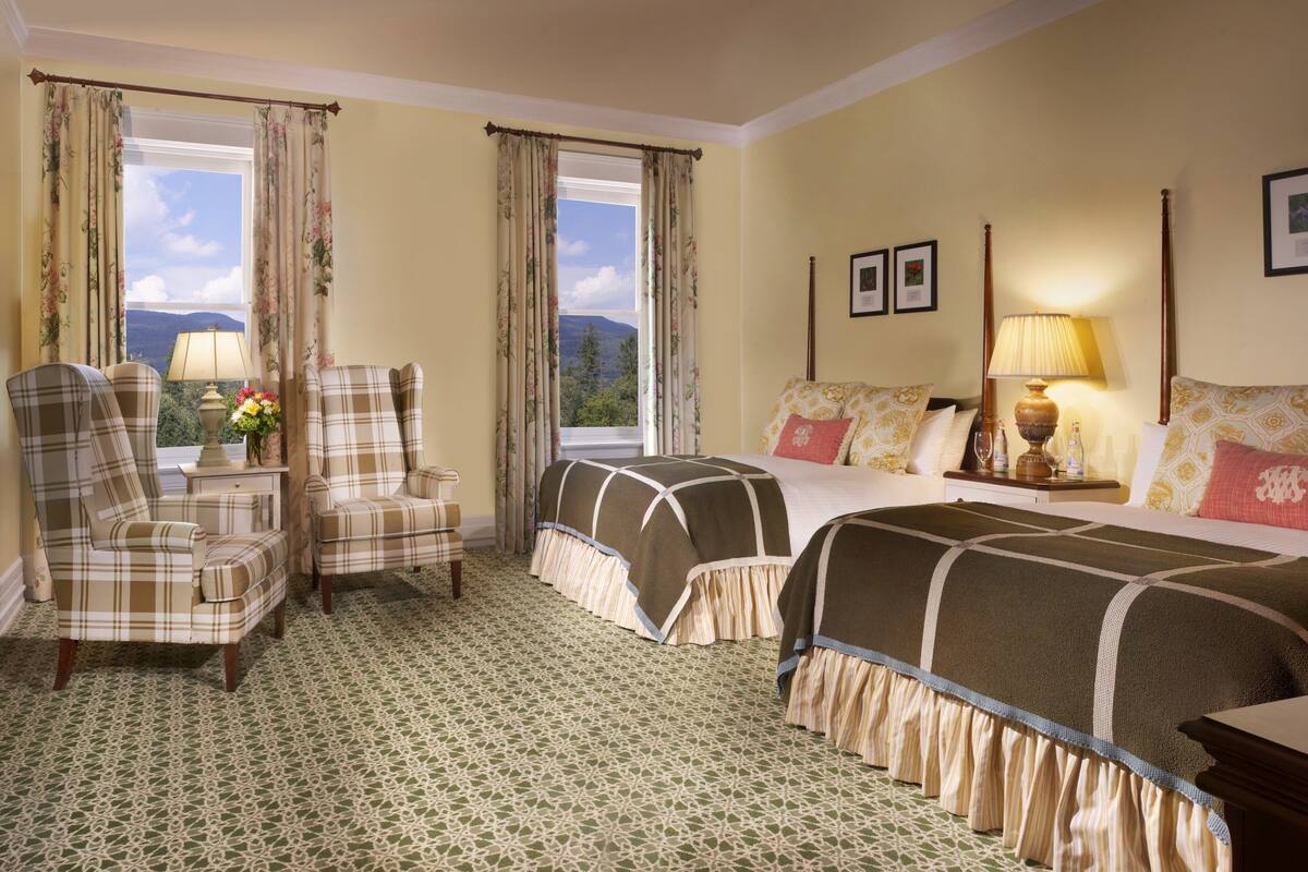 A hotel room with a large bed, two armchairs, a small table with a vase of flowers, and a view of mountains through the window.