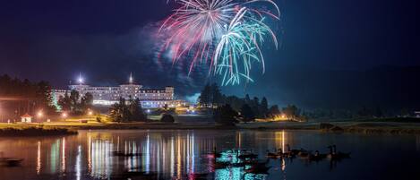 Un hotel amb el reflex de focs artificials en un llac, vaixells i un edifici ben il·luminat.