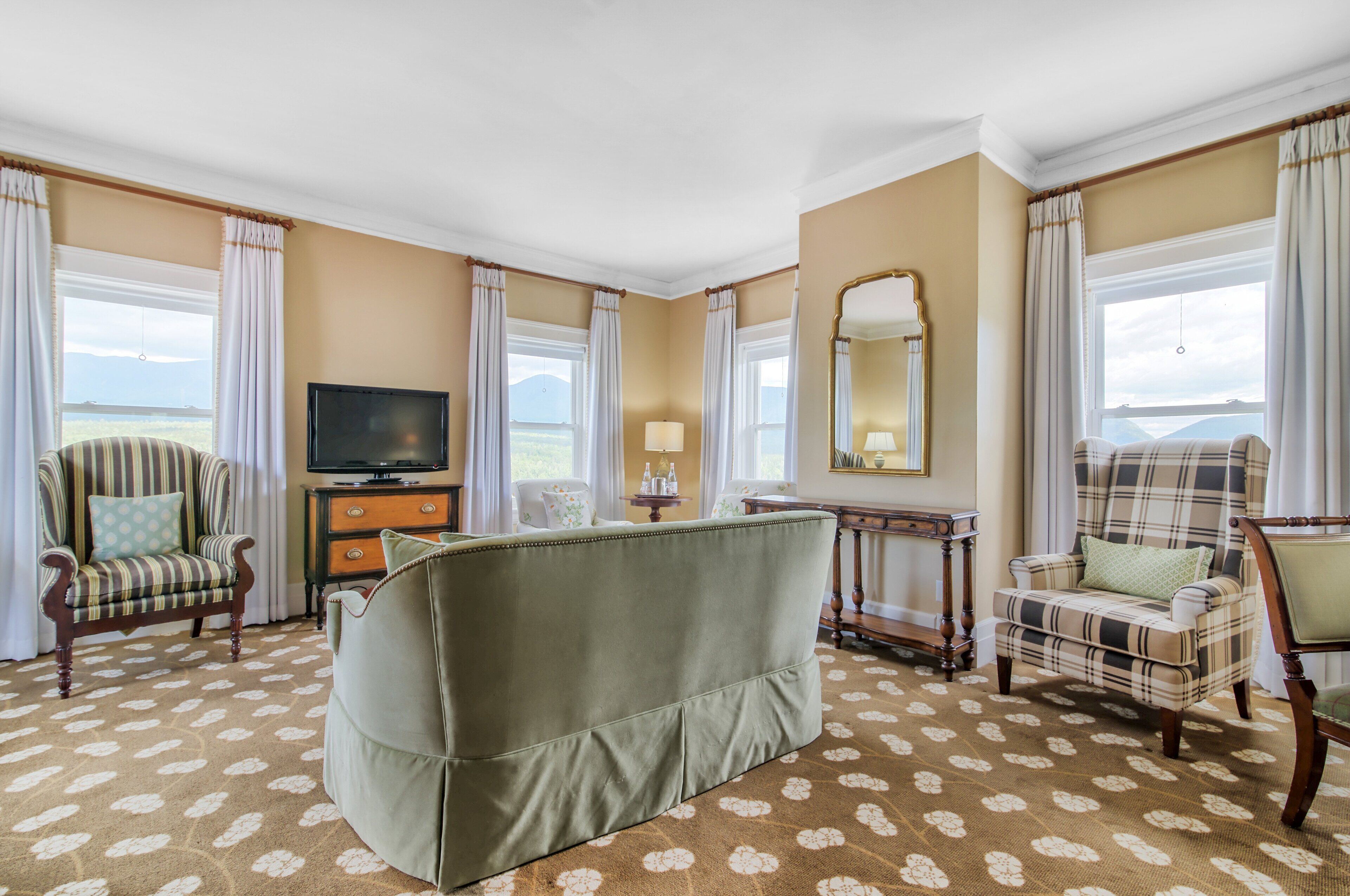 A hotel room with a large sofa, two armchairs, a TV, a mirror, and a view of the mountains.