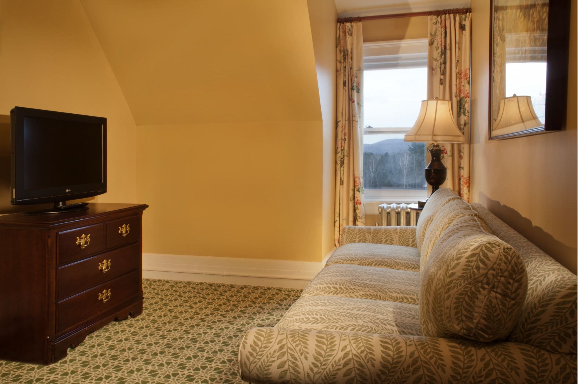 A hotel room with a television on a wooden dresser, a patterned sofa, a window with curtains, and a lamp.