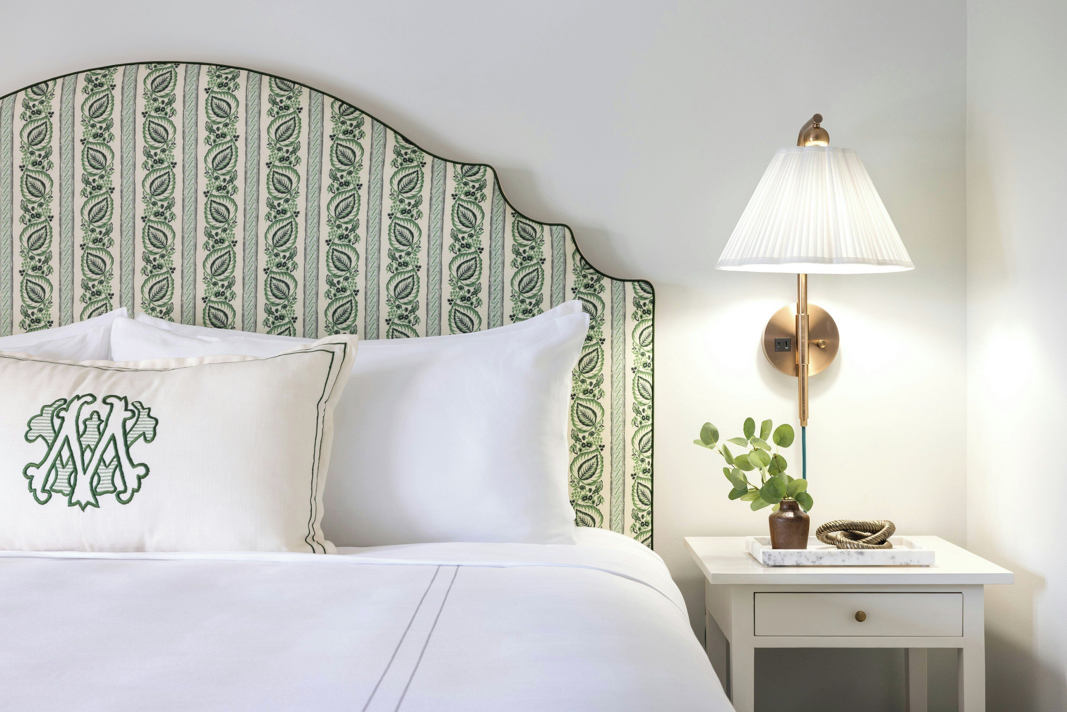 Una cama bien hecha con sábanas blancas, una almohada decorativa con un monograma, un cabecero estampado, una lámpara de noche y una planta pequeña en una mesita.
