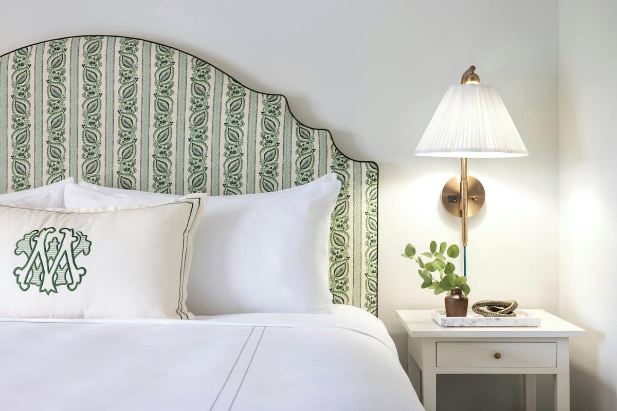 A neatly made bed with white linens, a decorative pillow with a monogram, a patterned headboard, a bedside lamp, and a small plant on a nightstand.