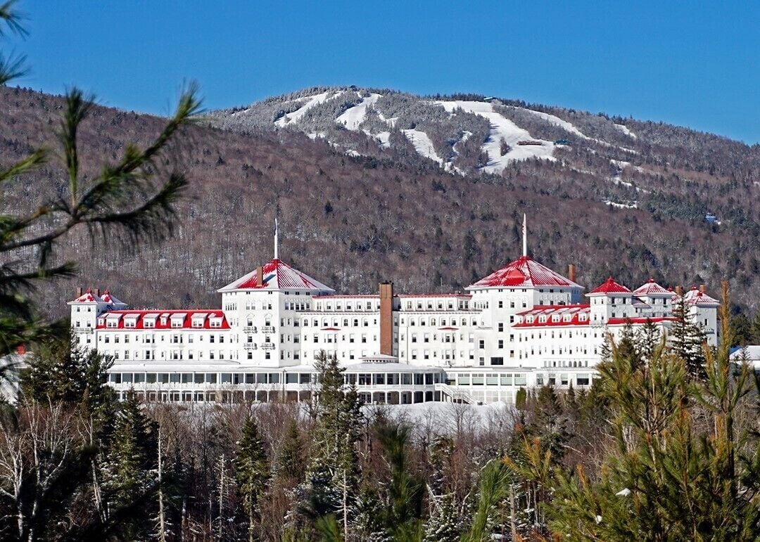 A large, multi-story hotel with red roofs and white walls, surrounded by trees and a mountain in the background.