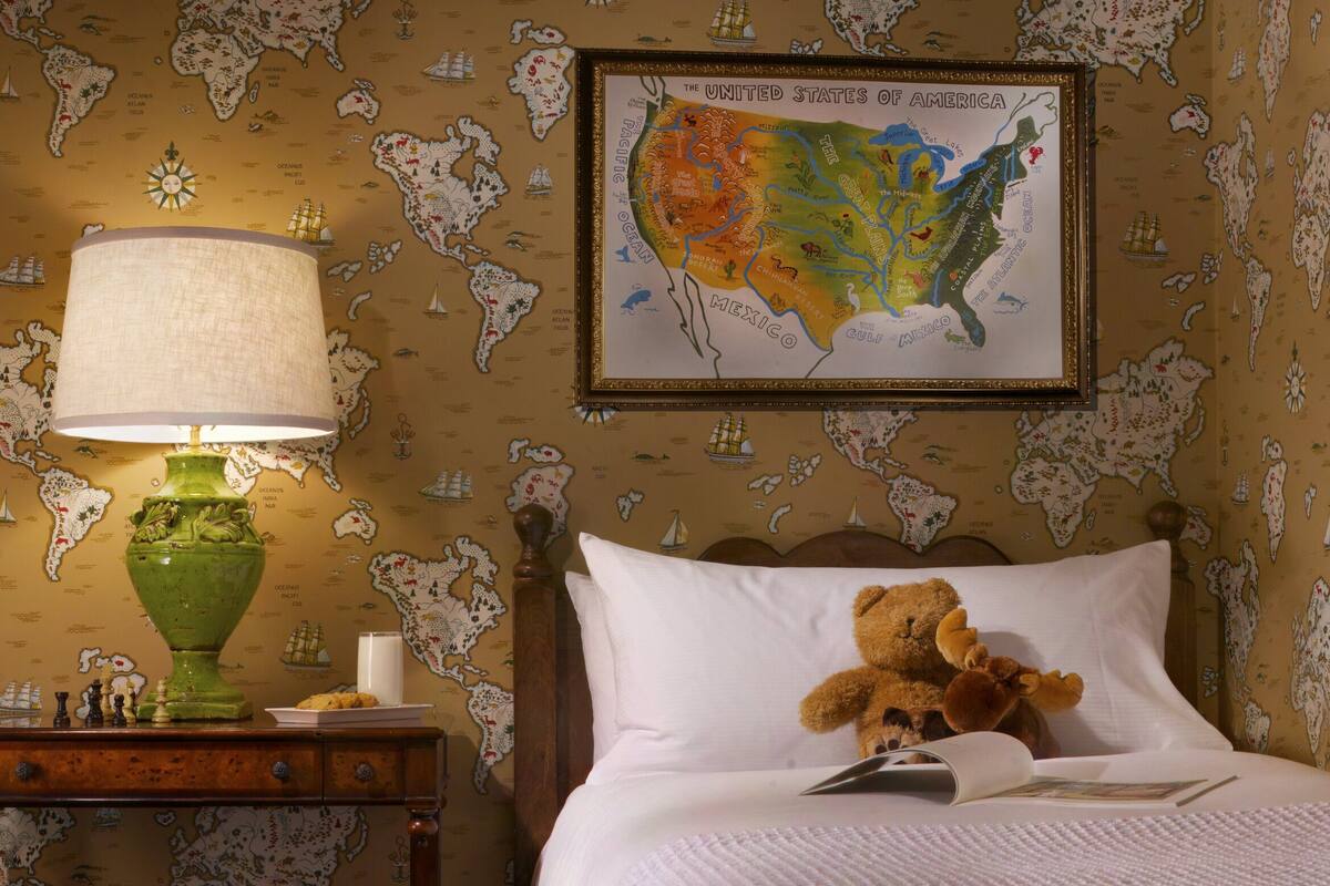 A hotel room with a bed, a nightstand with a lamp, a world map on the wall, and a teddy bear on the bed.