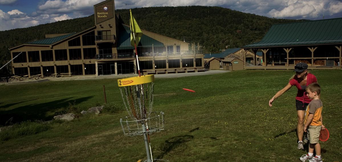 Frisbeegolfrata, jossa on selkeästi merkitty frisbeegolfteline ja lippu, vihreäkattoinen rakennus ja taustalla vuoristomaisema.