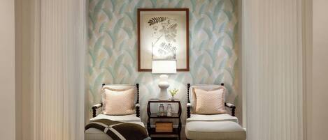 A room with two upholstered chairs, a small table with a lamp, and a framed botanical artwork on the wall.