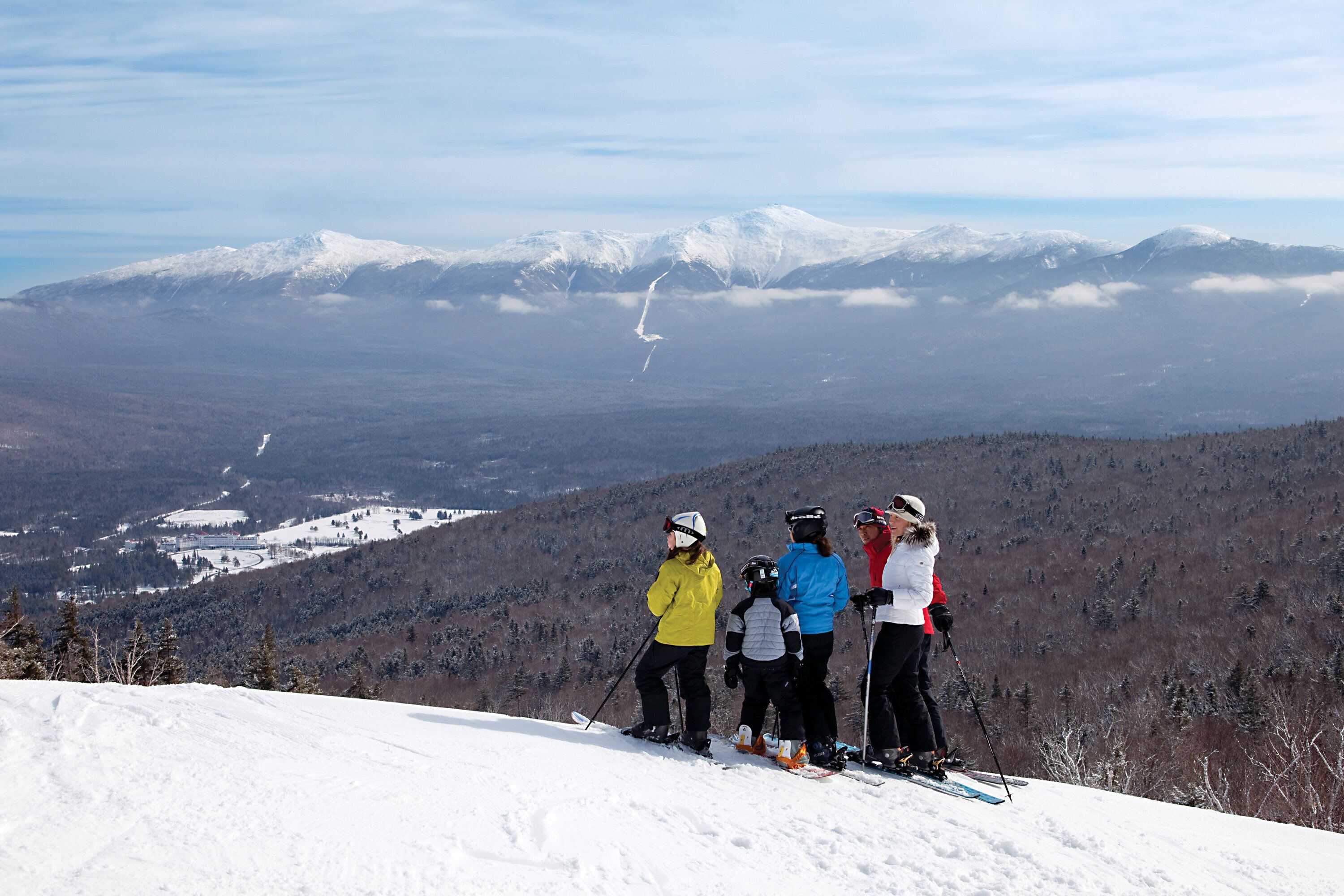 A group of skiers on a snowy mountain slope with a view of distant snow-covered peaks and a valley below.