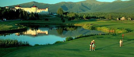 Um campo de golfe com um edifício grande ao fundo, um lago refletindo a paisagem e uma paisagem montanhosa.