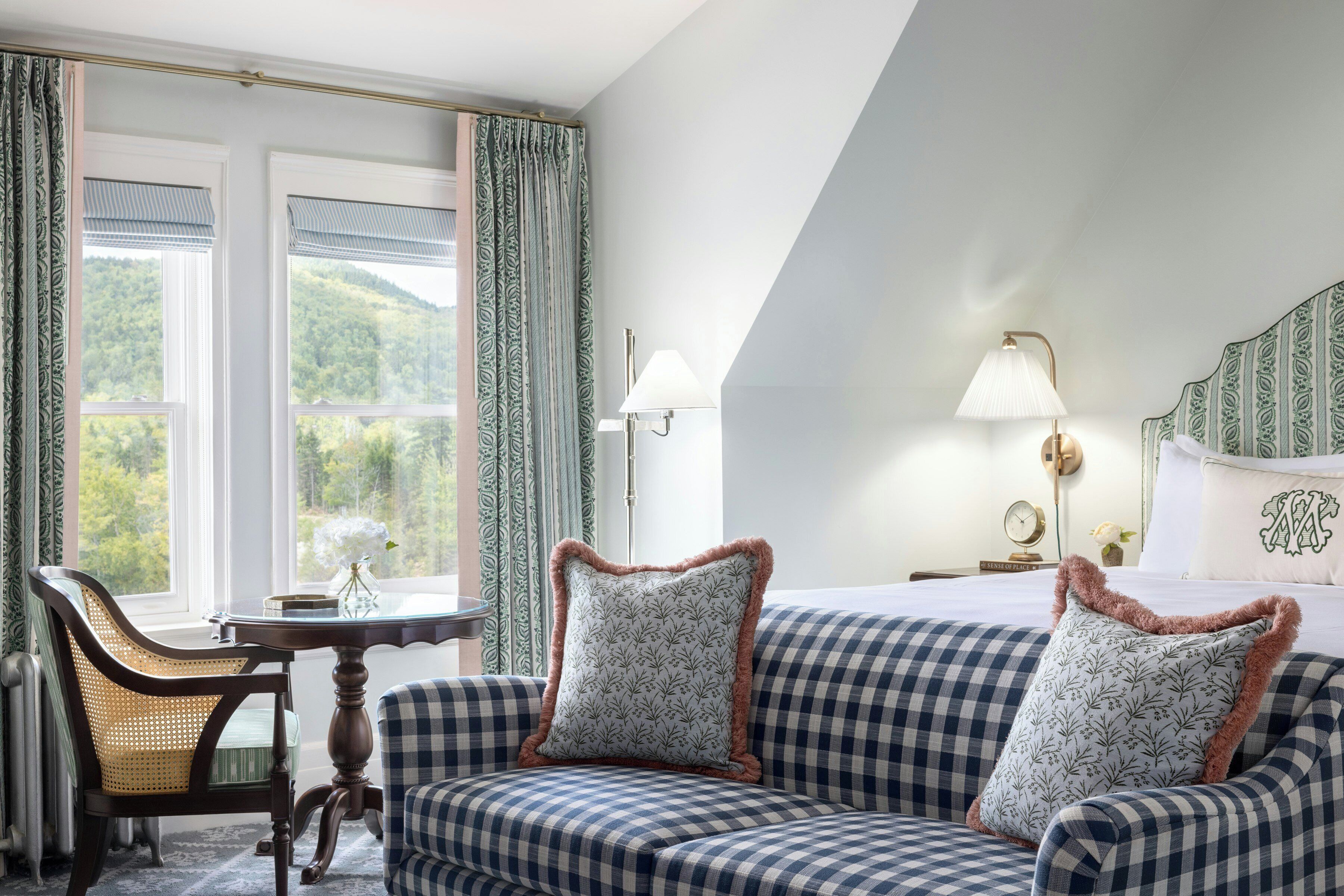A room with a checkered sofa, a table with a vase, a lamp, and a view of greenery through the windows.