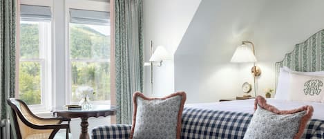 A room with a checkered sofa, a table with a vase, a lamp, and a view of greenery through the windows.