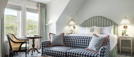A bedroom with a checkered sofa, a bed with a patterned headboard, two bedside lamps, a small table with a book, and a view of greenery through the window.