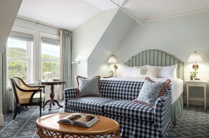 A bedroom with a checkered sofa, a bed with a patterned headboard, two bedside lamps, a small table with a book, and a view of greenery through the window.