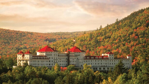 A large, multi-story building with red roofs, surrounded by autumn-colored trees on a hillside.