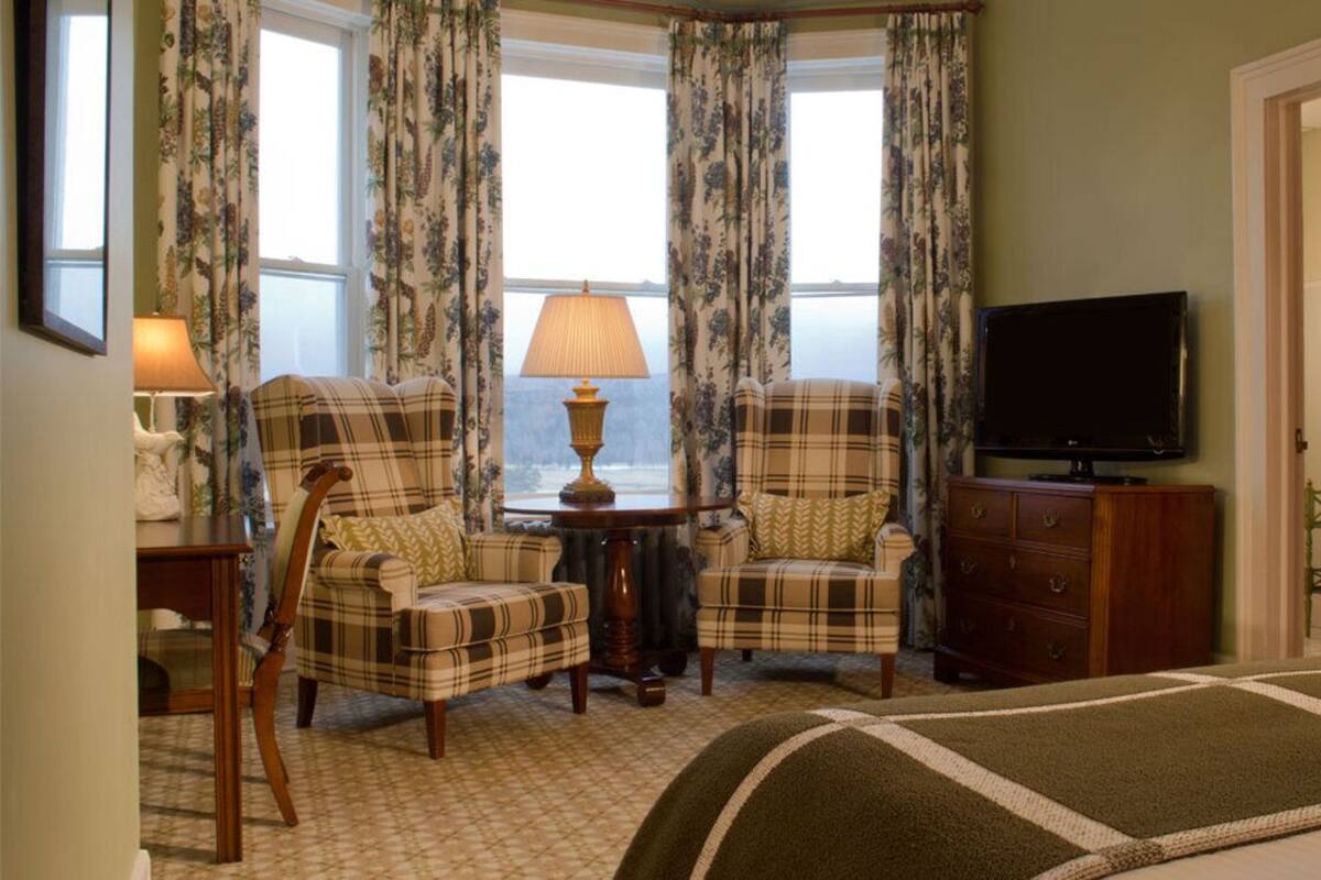 A hotel room with a bed, two plaid armchairs, a small table with a lamp, a television, and a view of the outdoors through large windows.