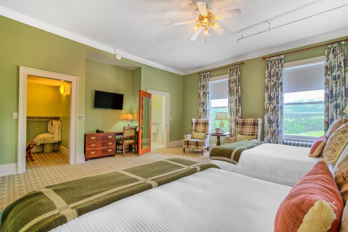 A hotel room with two beds, a desk, a chair, a television, and a view of the outdoors.
