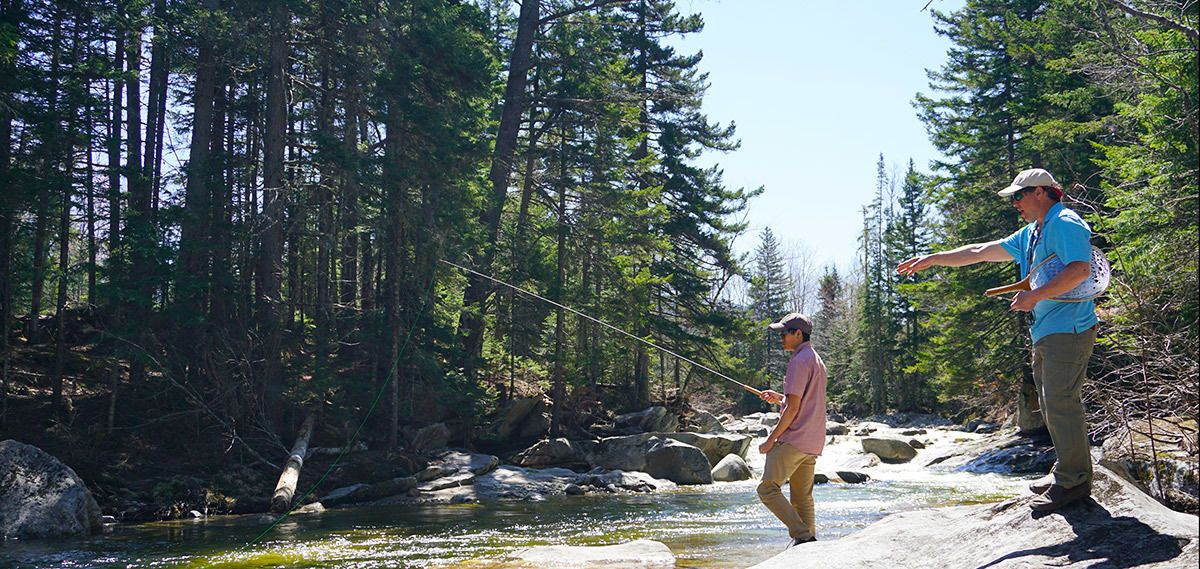 Two people fishing by a river in a forested area.