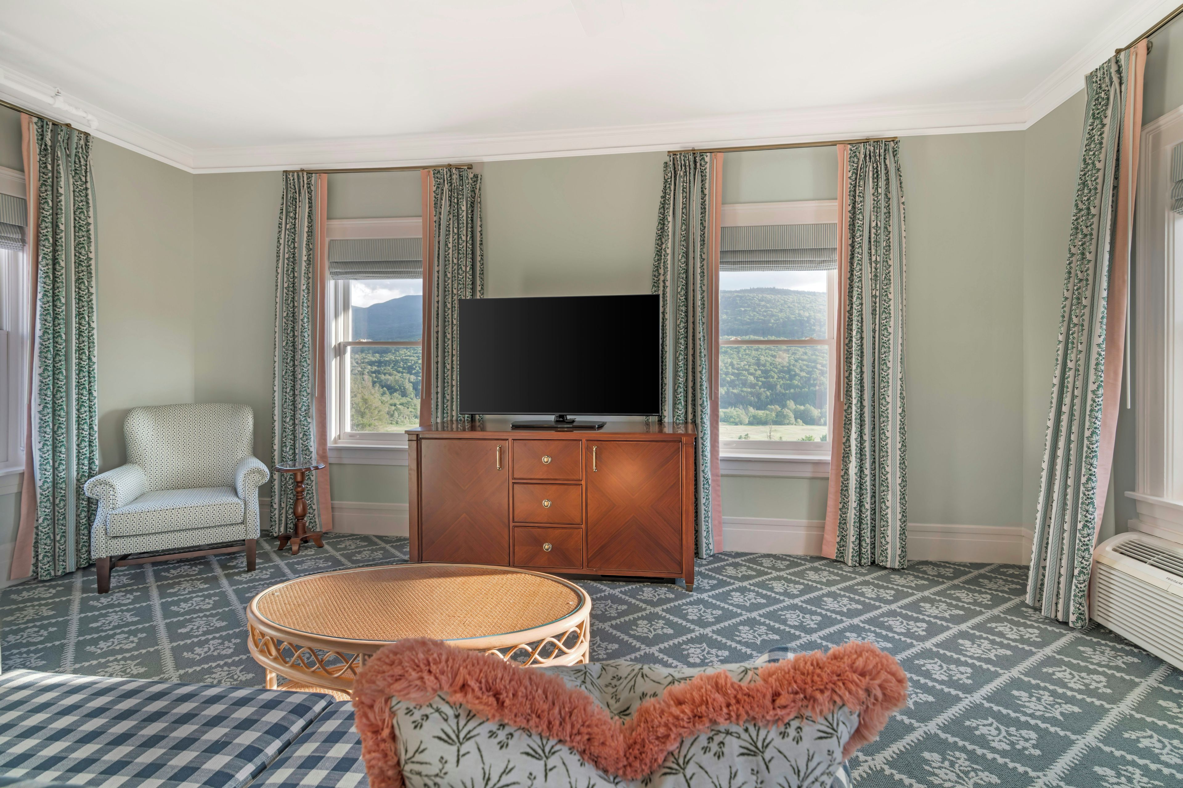 A room with a television on a wooden cabinet, a patterned armchair, a round wicker table, and a view of a mountainous landscape through the windows.