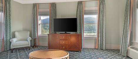 A room with a television on a wooden cabinet, a patterned armchair, a round wicker table, and a view of a mountainous landscape through the windows.