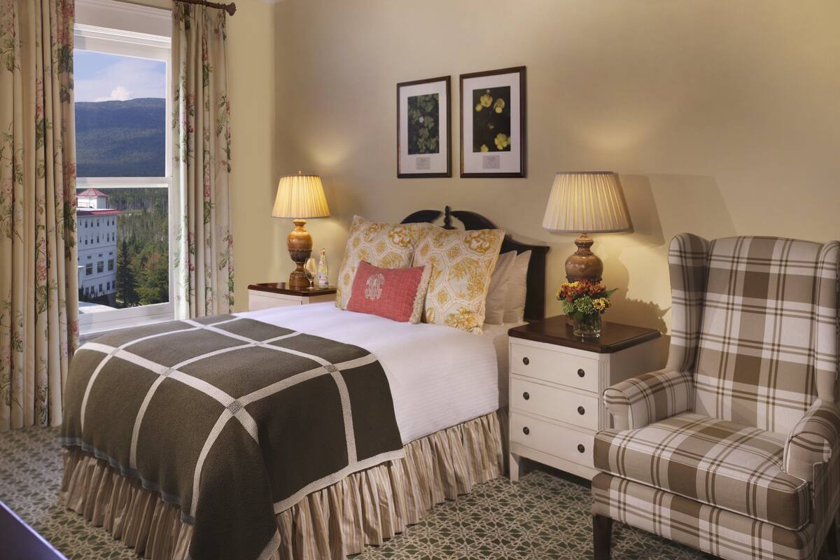 A hotel room with a bed, two bedside lamps, a plaid armchair, a nightstand, and a window with curtains.