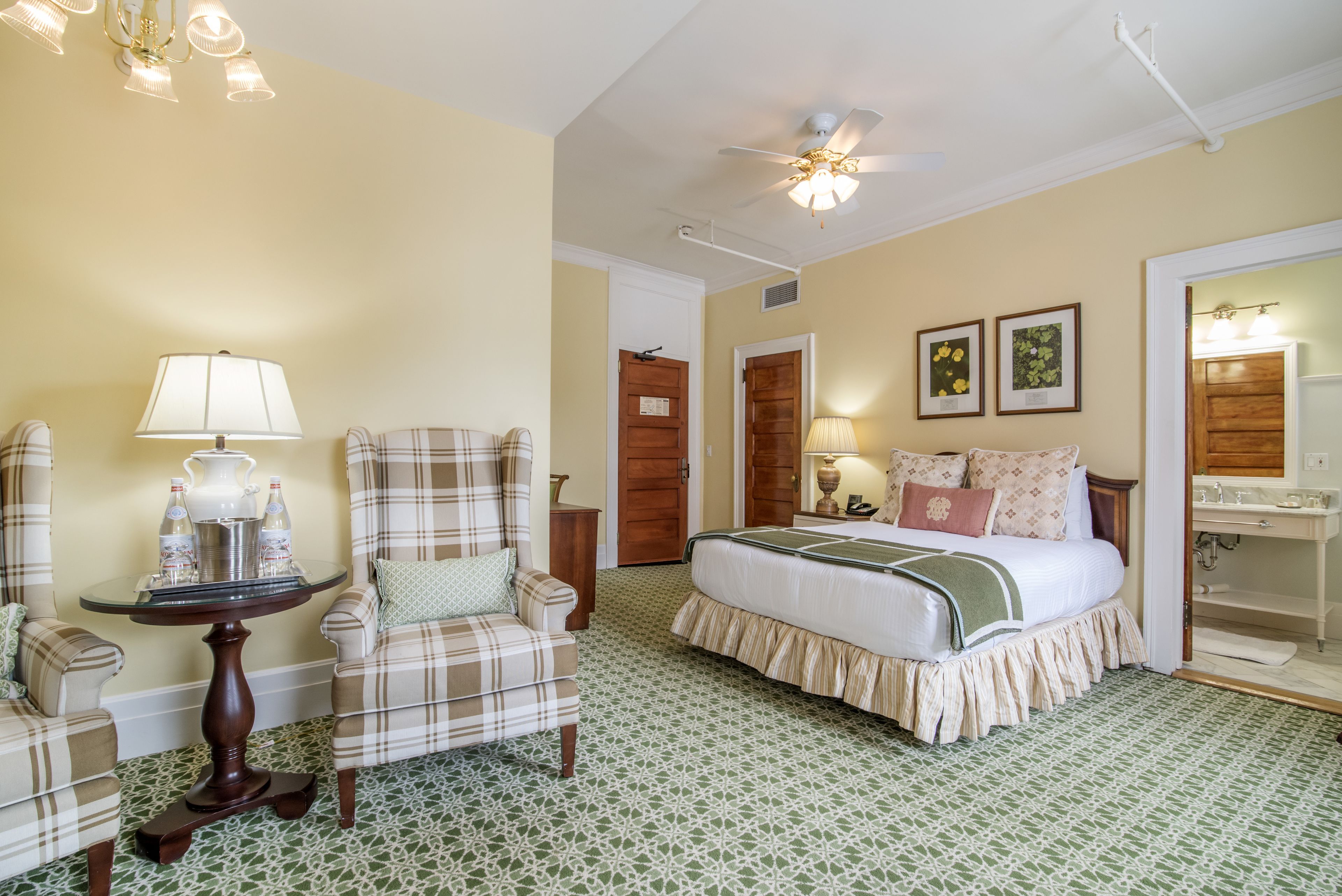 A bedroom with a bed, a chair, a table with a lamp, and a ceiling fan.