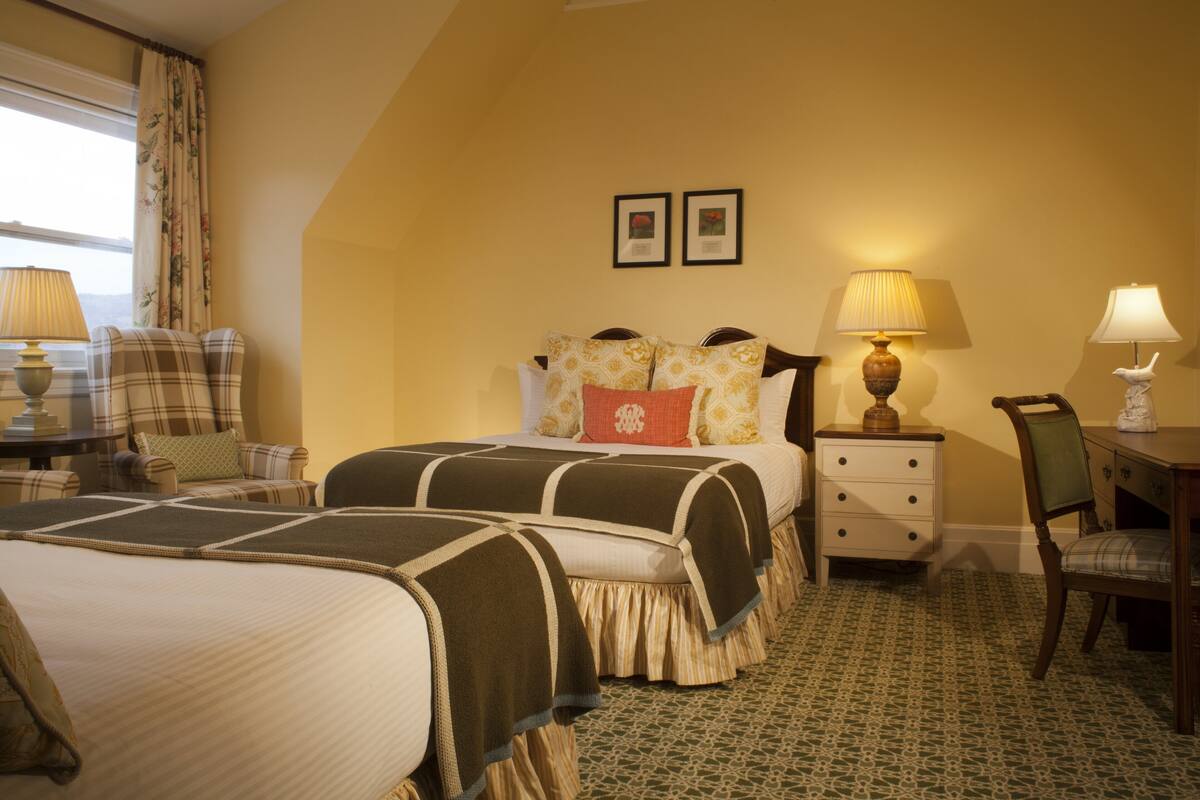 A hotel room with two beds, a desk, and a chair. There are two lamps on the desk and a window with curtains.