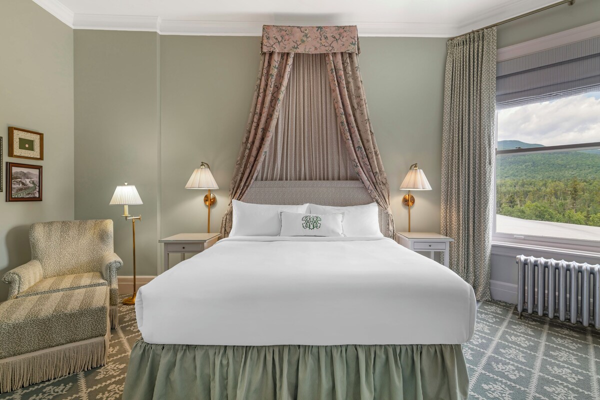 Habitación de hotel con una cama grande, dos mesitas de noche con lámparas, una silla y vistas al exterior.