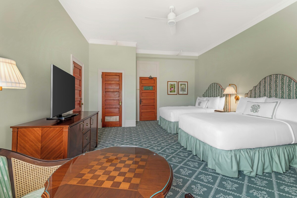 Une chambre d’hôtel équipée d’une télévision, d’un bureau, d’une chaise, d’un lit avec deux oreillers, d’une table de chevet avec une lampe et d’un ventilateur de plafond.