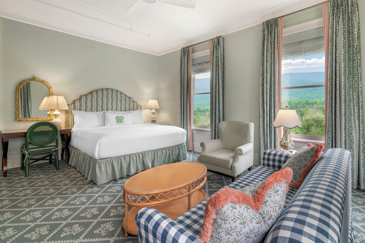 Een hotelkamer met een bed, twee stoelen, een bank, een tafel, een spiegel en uitzicht op het omliggende landschap.