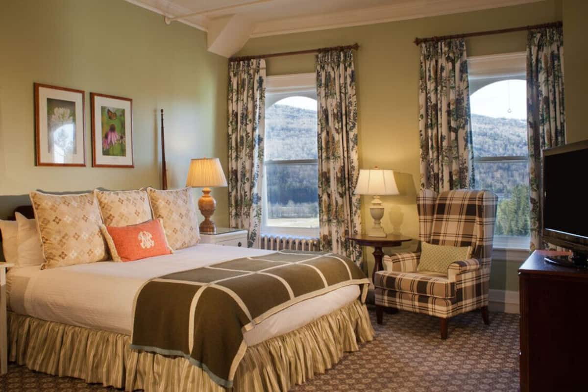 A bedroom with a large bed, two bedside tables with lamps, a chair, a television, and a window with curtains.