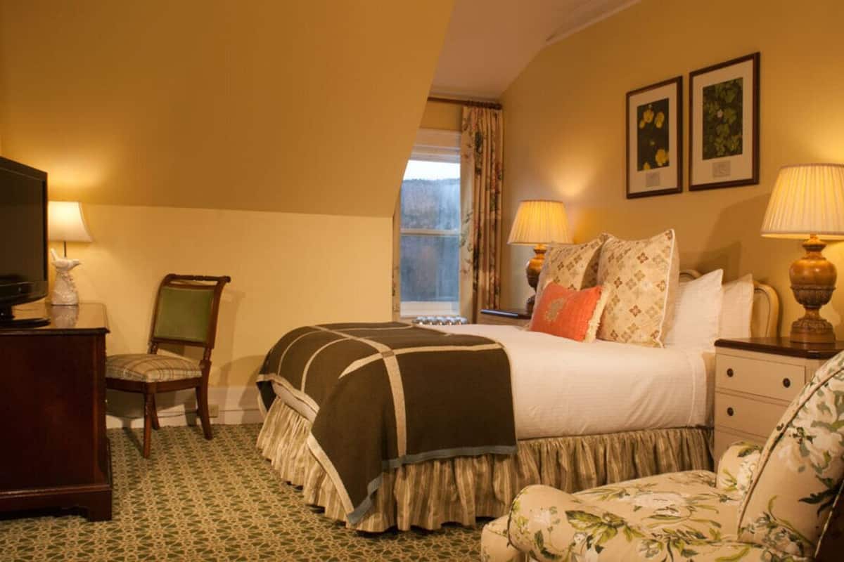 A hotel room with a bed, bedside lamps, a chair, a desk with a television, and a window with curtains.