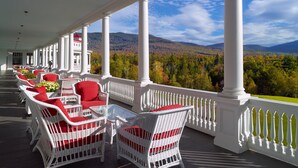 A covered outdoor seating area with white wicker furniture and red cushions, overlooking a mountainous landscape.