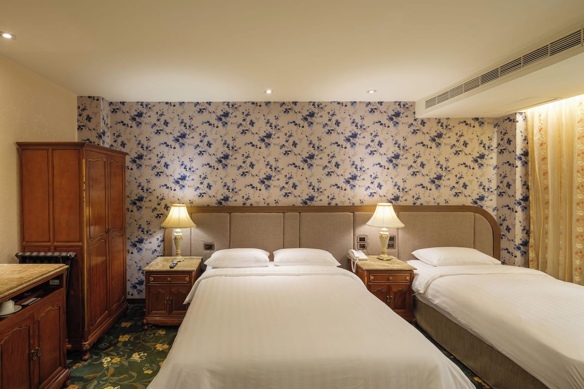 A hotel room with two beds, a wooden wardrobe, a nightstand with a lamp, and floral wallpaper.