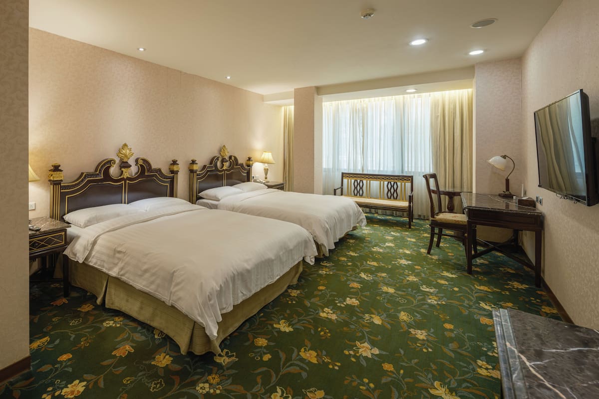 A hotel room with two beds, a desk, a chair, a TV, and a window with curtains.