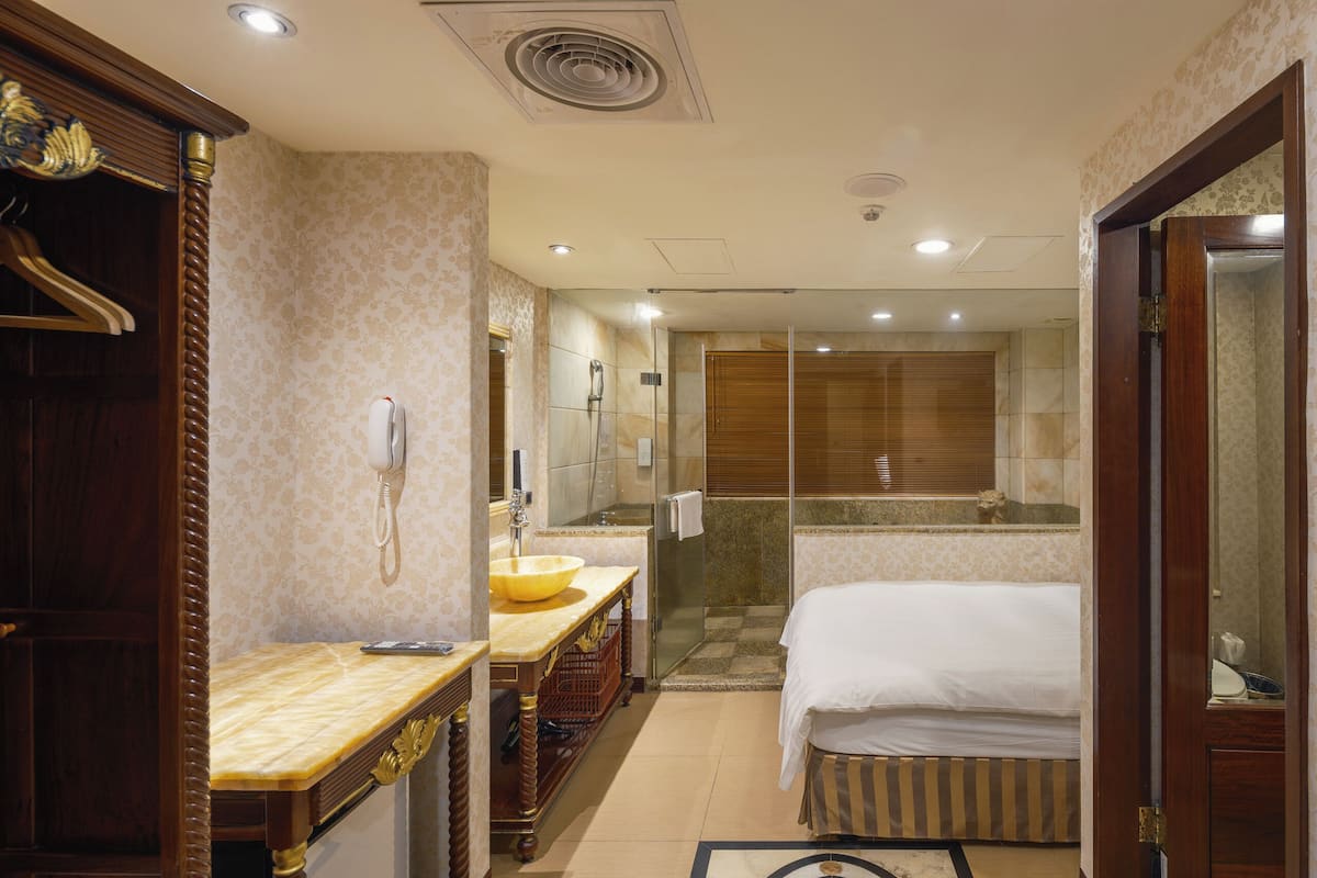 A hotel room with a bed, a vanity with a sink, a shower, and a wooden wardrobe.