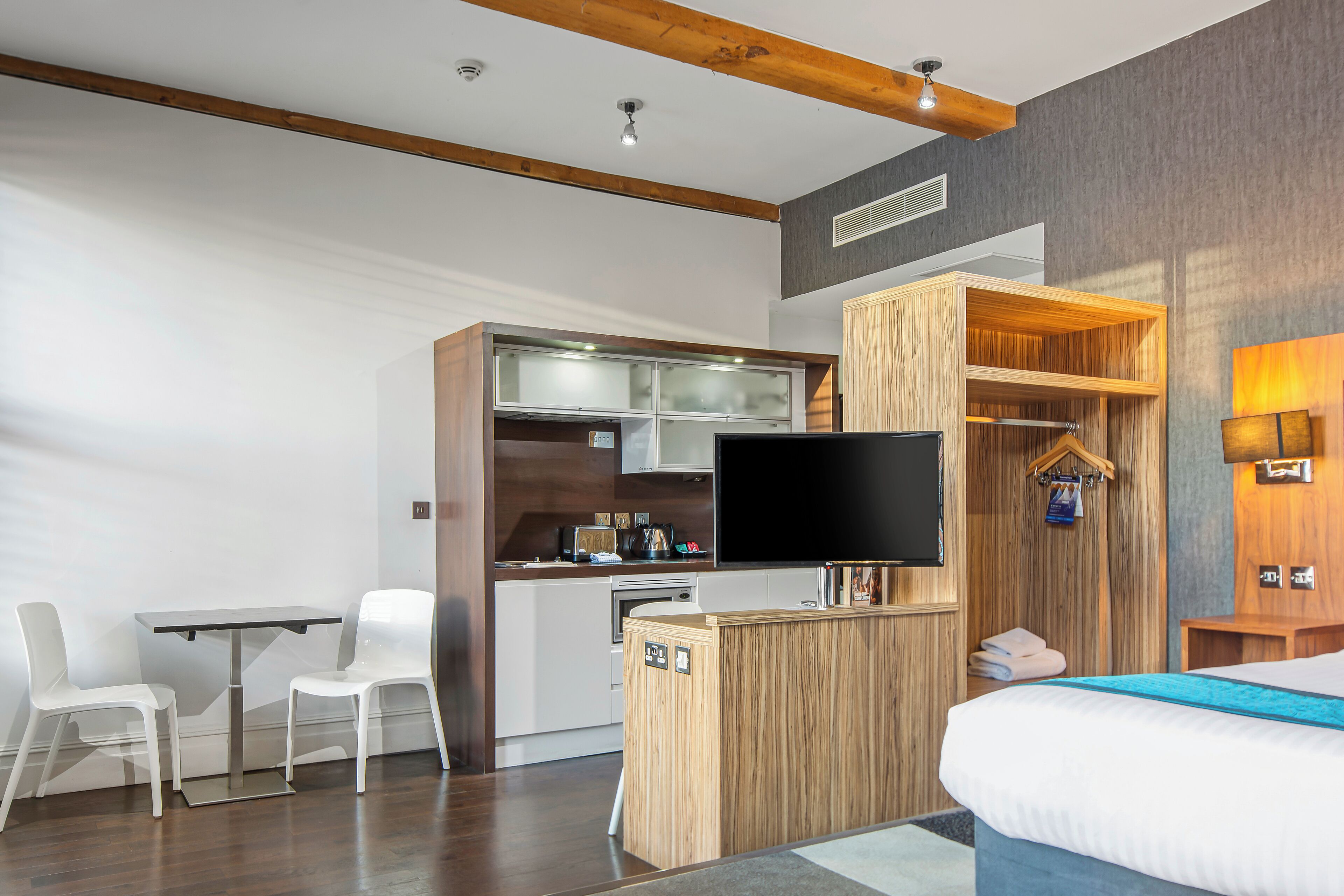 Et moderne hotelværelse med skrivebord i træ, fladskærms-tv, seng med blå detaljer og indbygget garderobeskab.