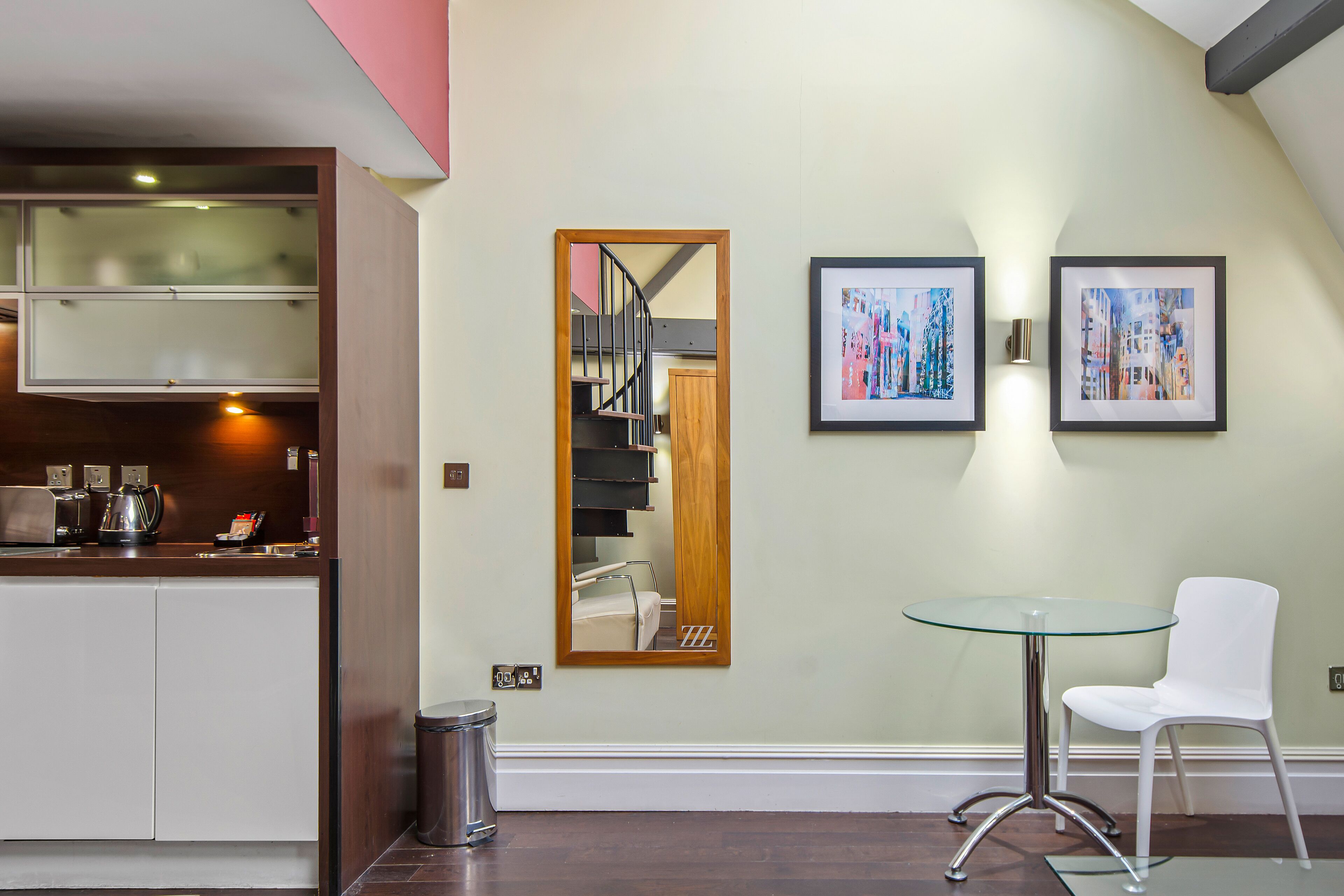 A modern kitchen with a built-in stove, a coffee maker, and a trash can. There is a glass table with a white chair and two framed artworks on the wall.