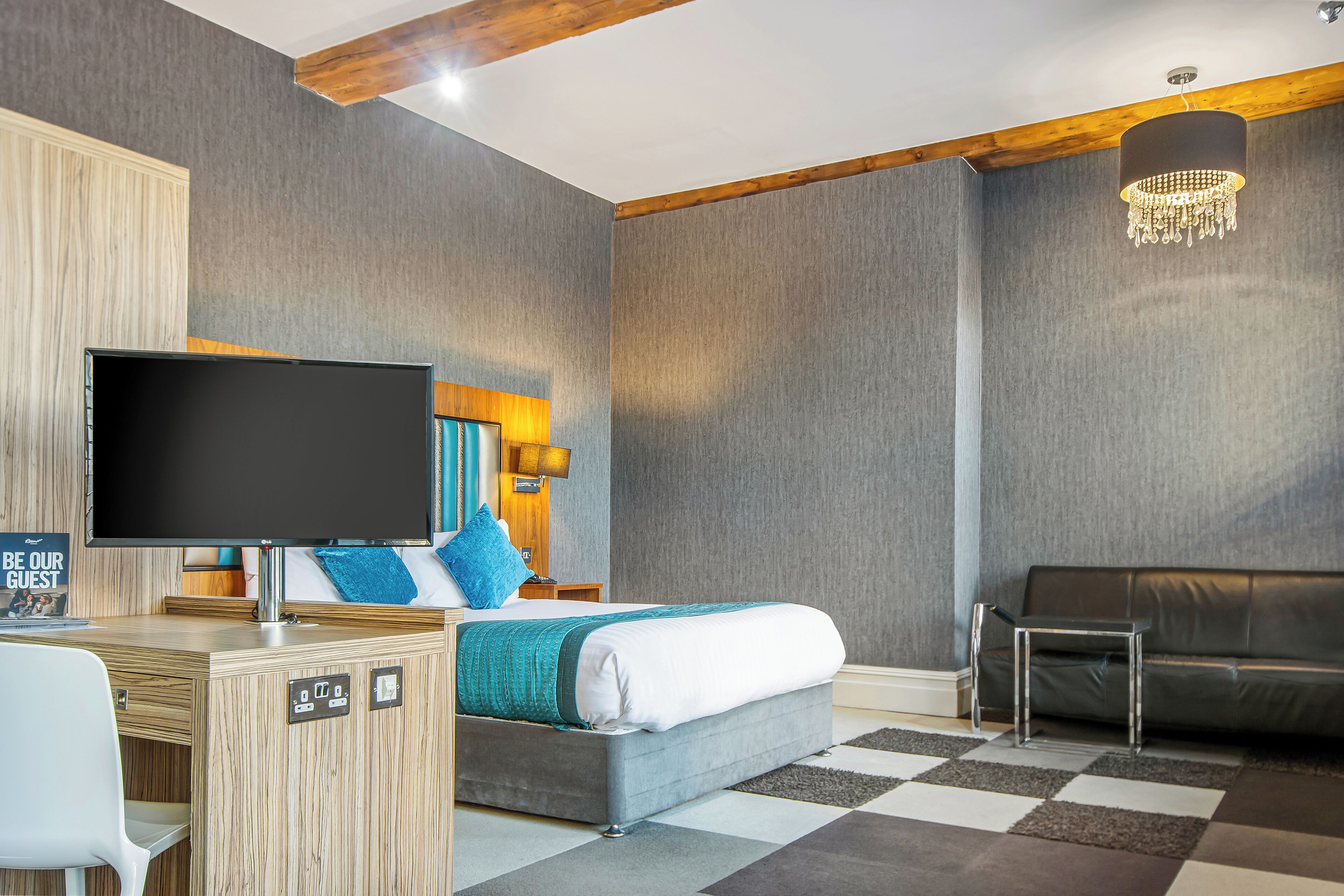 A modern hotel room with a large bed, a desk with a flat-screen TV, a sofa, and a chandelier.