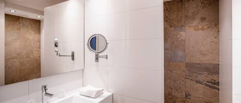 Un baño moderno con lavamanos blanco, espejo y zona de ducha con azulejos color beige.